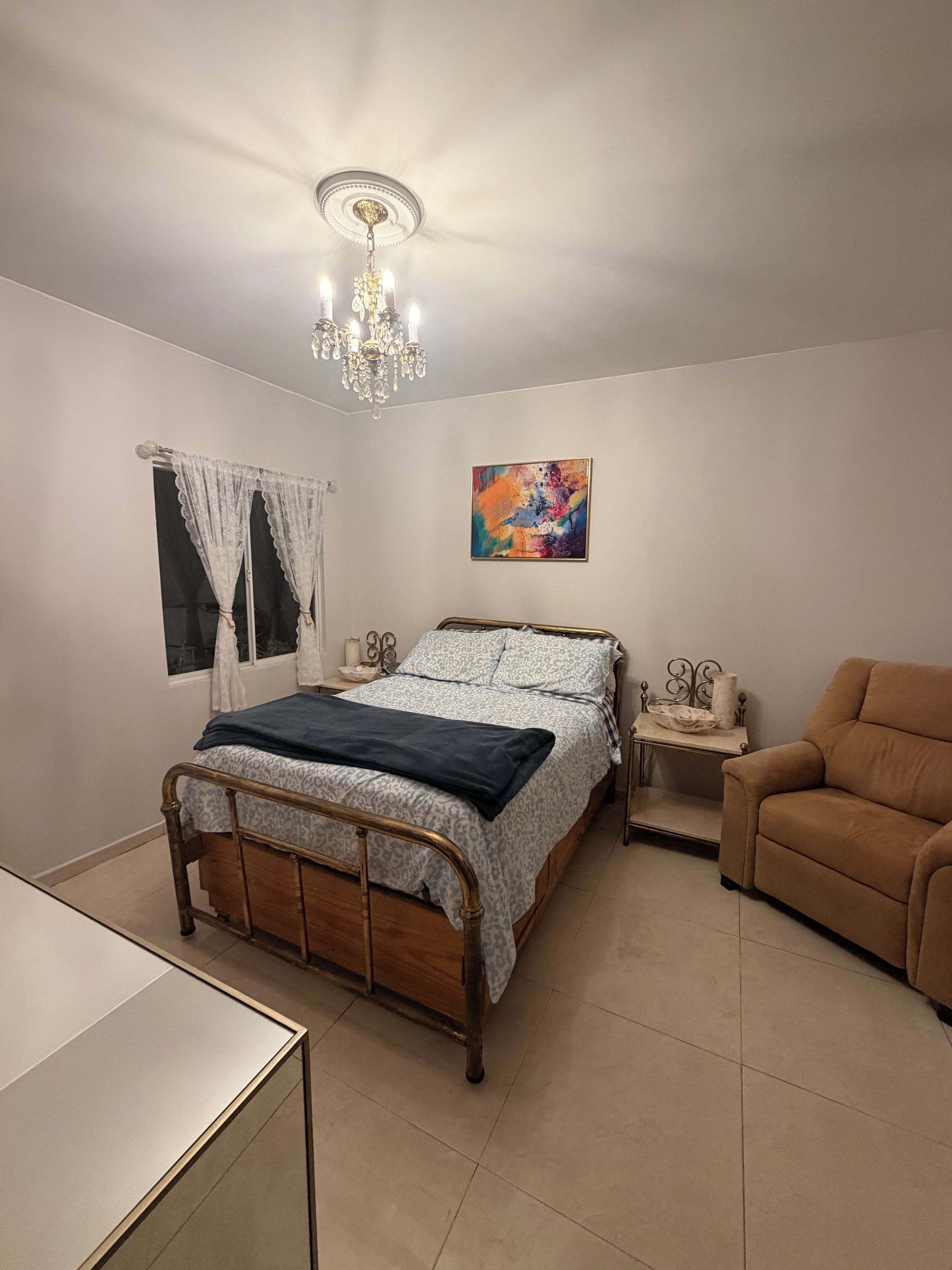 Une chambre à coucher comprenant un lit, une table de chevet, une chaise, une fenêtre avec des rideaux, un tableau et un lustre.