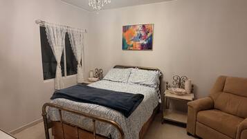 Une chambre à coucher comprenant un lit, une table de chevet, une chaise, une fenêtre avec des rideaux, un tableau et un lustre.