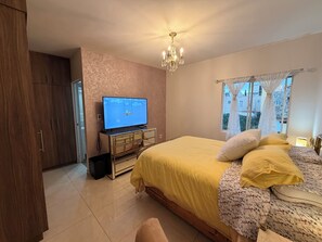 A bedroom with a bed, a television on a dresser, a chandelier, and a window with a view of buildings.