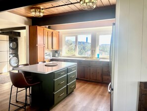 A modern kitchen with wooden cabinets, a central island, a built-in oven, and a large window offering a view of the outdoors. - Vista Grande-2 Hot Tubs! 3 Large Decks! Arcade! (Ruidoso)
