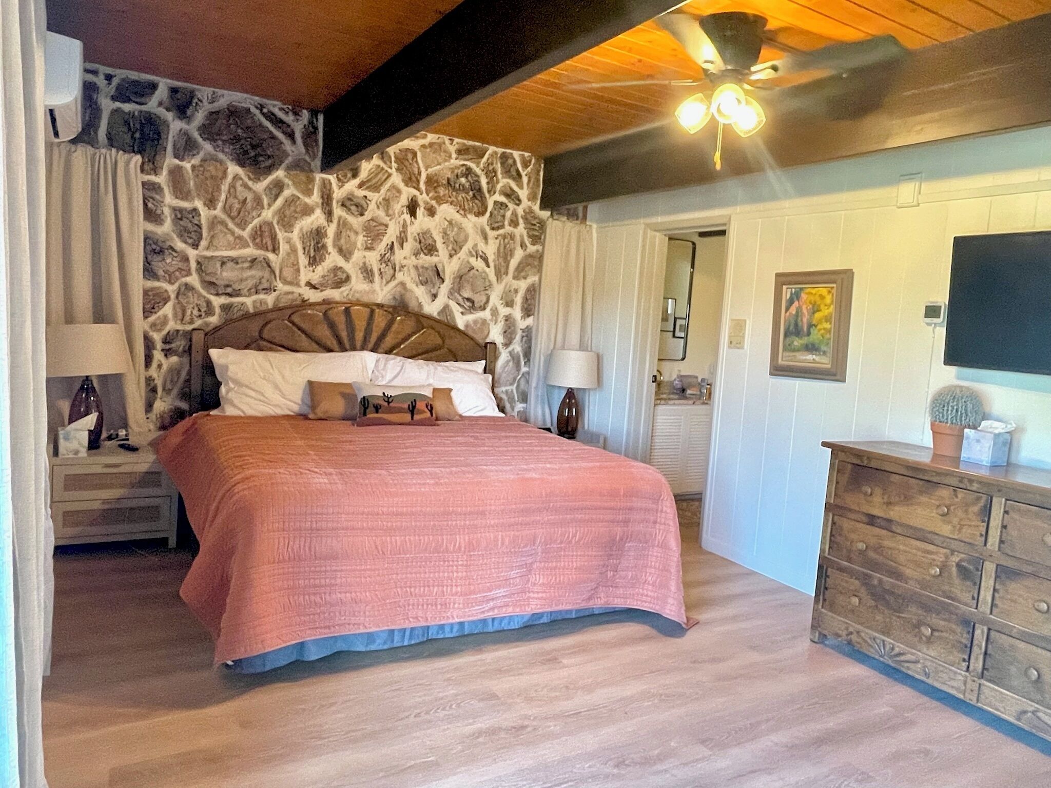 A bedroom with a stone wall, a bed with a wooden headboard, a dresser, and a ceiling fan.
