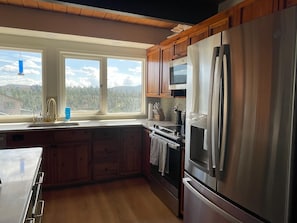 A modern kitchen with wooden cabinets, stainless steel appliances, a large window, and a view of a forested area. - Vista Grande-2 Hot Tubs! 3 Large Decks! Arcade! (Ruidoso)