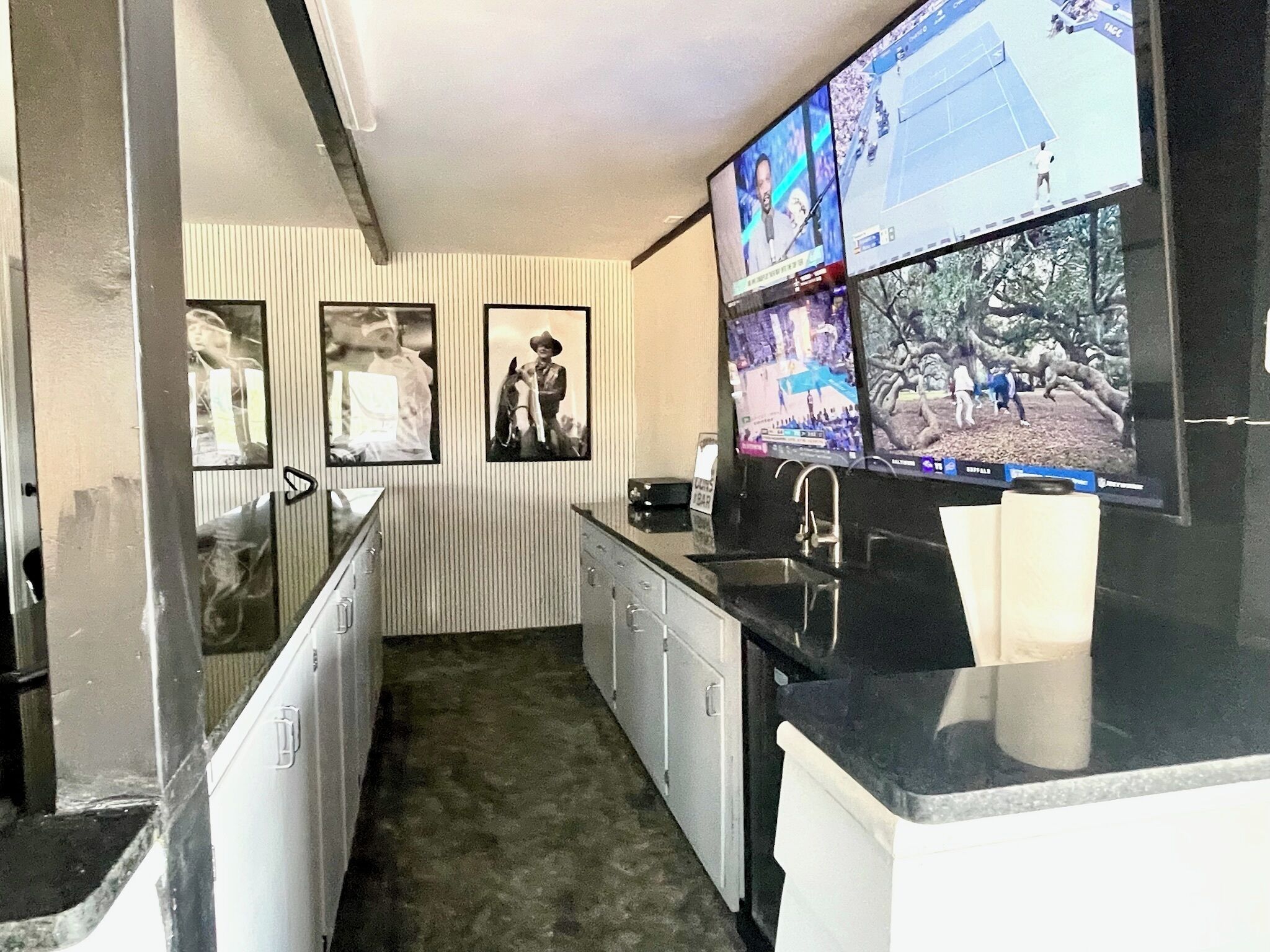 A kitchen with a large flat-screen TV mounted above the counter, a sink, and a refrigerator. There are framed pictures on the wall.