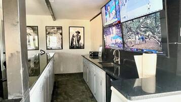 A kitchen with a large flat-screen TV mounted above the counter, a sink, and a refrigerator. There are framed pictures on the wall.