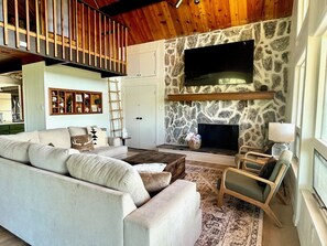 A living room with a stone fireplace, a sofa, a coffee table, and a television. - Vista Grande-2 Hot Tubs! 3 Large Decks! Arcade! (Ruidoso)