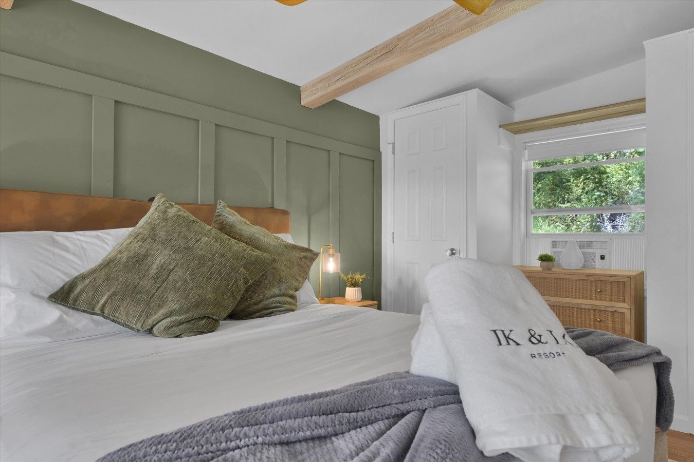 A neatly made bed with pillows, a bedside lamp, and a window with a view of greenery.