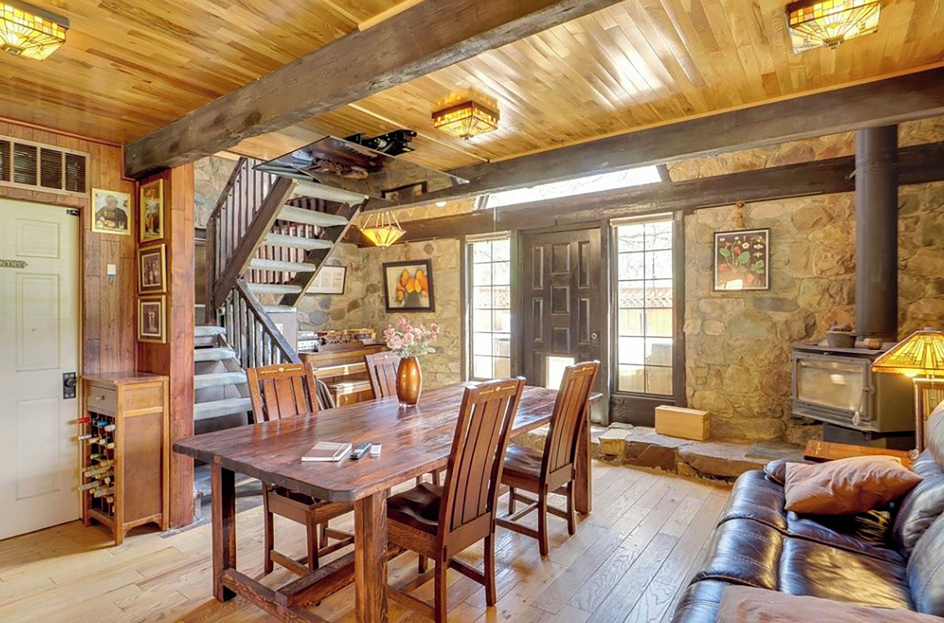 A rustic interior with a wooden dining table, chairs, a staircase, stone walls, and a fireplace.