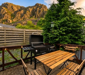 An outdoor patio with a barbecue grill, wooden table, and benches, surrounded by a wooden fence and greenery. - Mountain Backdrop Miners Cottage with Pizza Oven (Blaenau Ffestiniog)