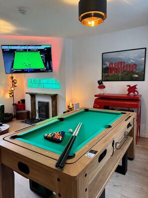 Een pooltafel met een neonbord, een televisie waarop een wedstrijd wordt vertoond, en een ingelijst beeld aan de muur.