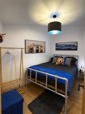 A bedroom with a bed, a hanging light fixture, a wall-mounted picture, a bedside table, and a small lamp. - Mountain Backdrop Miners Cottage with Pizza Oven (Blaenau Ffestiniog)