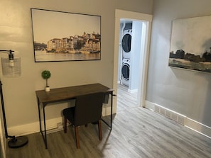 A room with a desk, chair, and two paintings. There is a door leading to a laundry area with washing machines.
