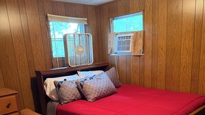 Quarto com parede revestida de madeira, cama com roupa de cama vermelha, mesa de cabeceira, cadeira e dois ventiladores fixados na parede.