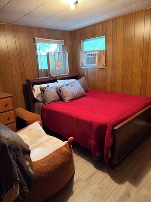 Ein Schlafzimmer mit einer holzverkleideten Wand, einem Bett mit roter Bettwäsche, einem Nachttisch, einem Stuhl und zwei Wandventilatoren.