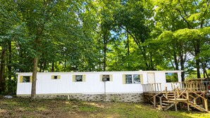Uma casa móvel com base de tijolos, cercada por árvores e um caminho de cascalho.