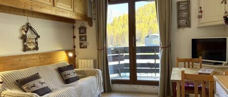 Una sala de estar acogedora con sofá, mesa de comedor y televisor. Se aprecia un ventanal con vistas a árboles y una montaña.