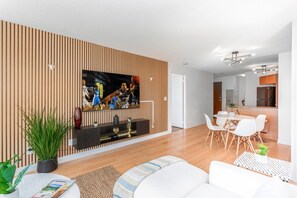 Ein modernes Wohnzimmer mit einem Flachbildfernseher, weißen Sofas, einem Essbereich mit Tisch und Stühlen sowie einer Küche im Hintergrund.