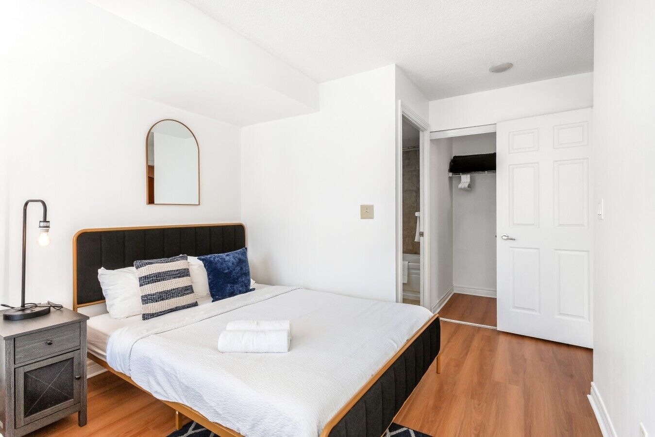Um quarto com cama, mesa de cabeceira, abajur e guarda-roupa.