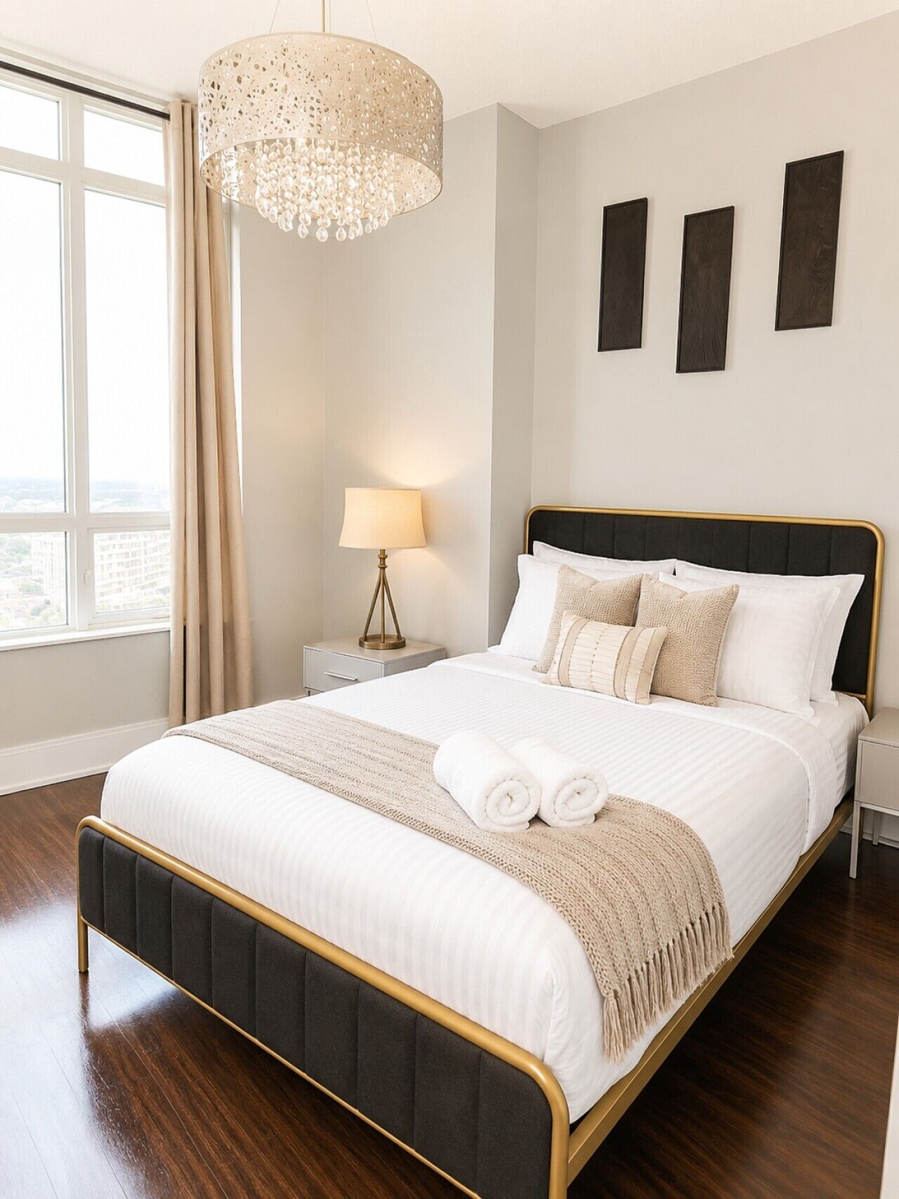 A modern bedroom with a large bed, a chandelier, a nightstand with a lamp, and a view of the city through the window.