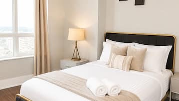 A modern bedroom with a large bed, a chandelier, a nightstand with a lamp, and a view of the city through the window.