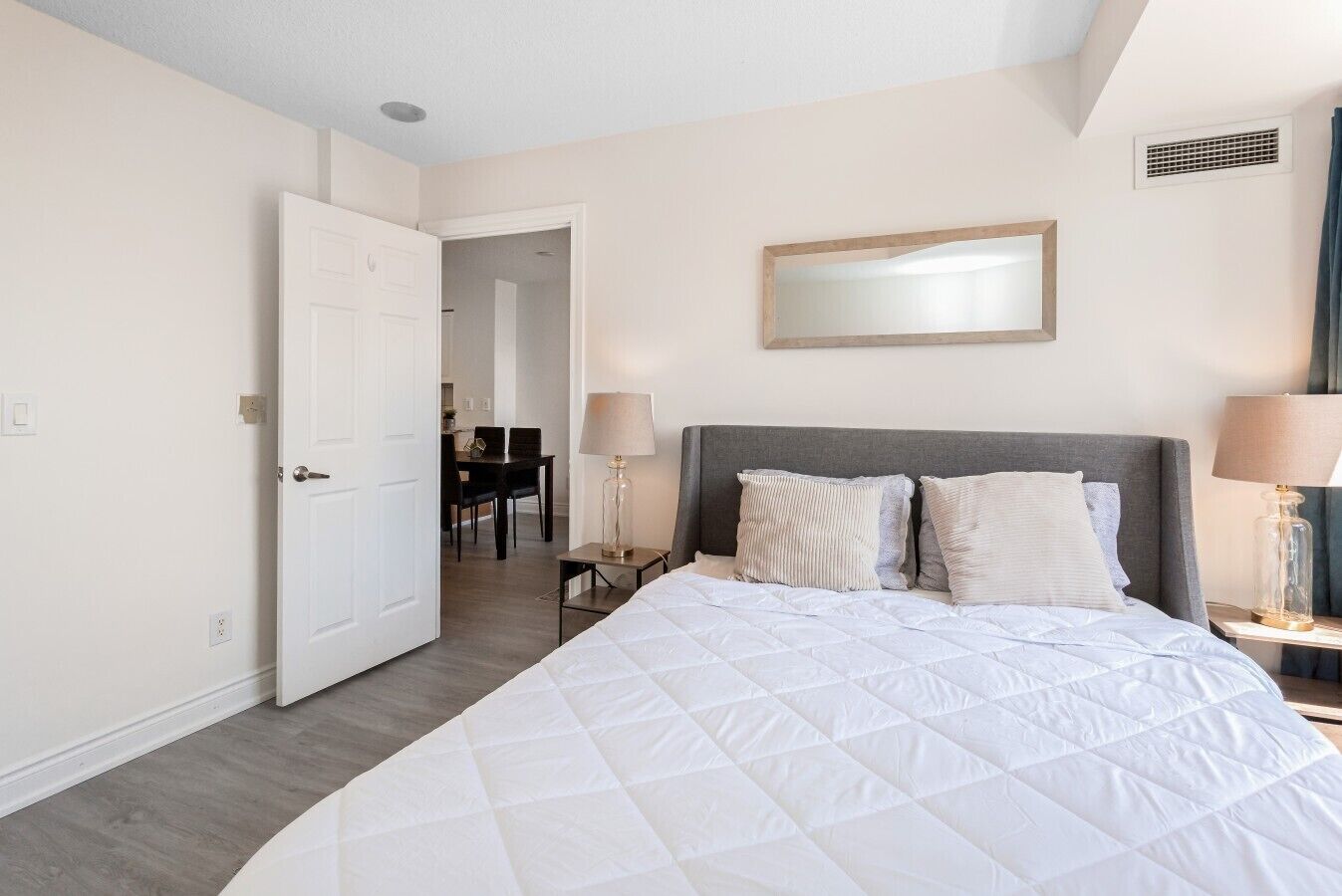A hotel room with a large bed, a bedside table with a lamp, a door leading to a dining area, and a wall-mounted mirror.