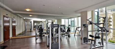 A gym with various exercise machines, wooden flooring, and large windows offering views of the city.