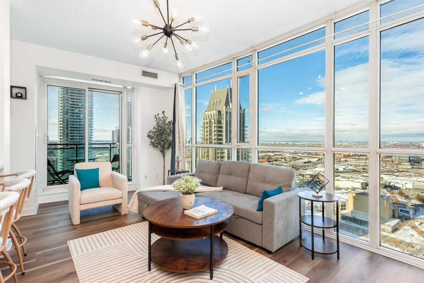 A spacious living room with a large window, a modern chandelier, and a view of the cityscape.