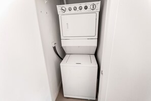 A compact, white laundry room with a front-loading washer and dryer, a visible power outlet, and a door leading to another room. - Sky-High 2BR/2BA Condo | Best City Views+Parking (Mississauga)