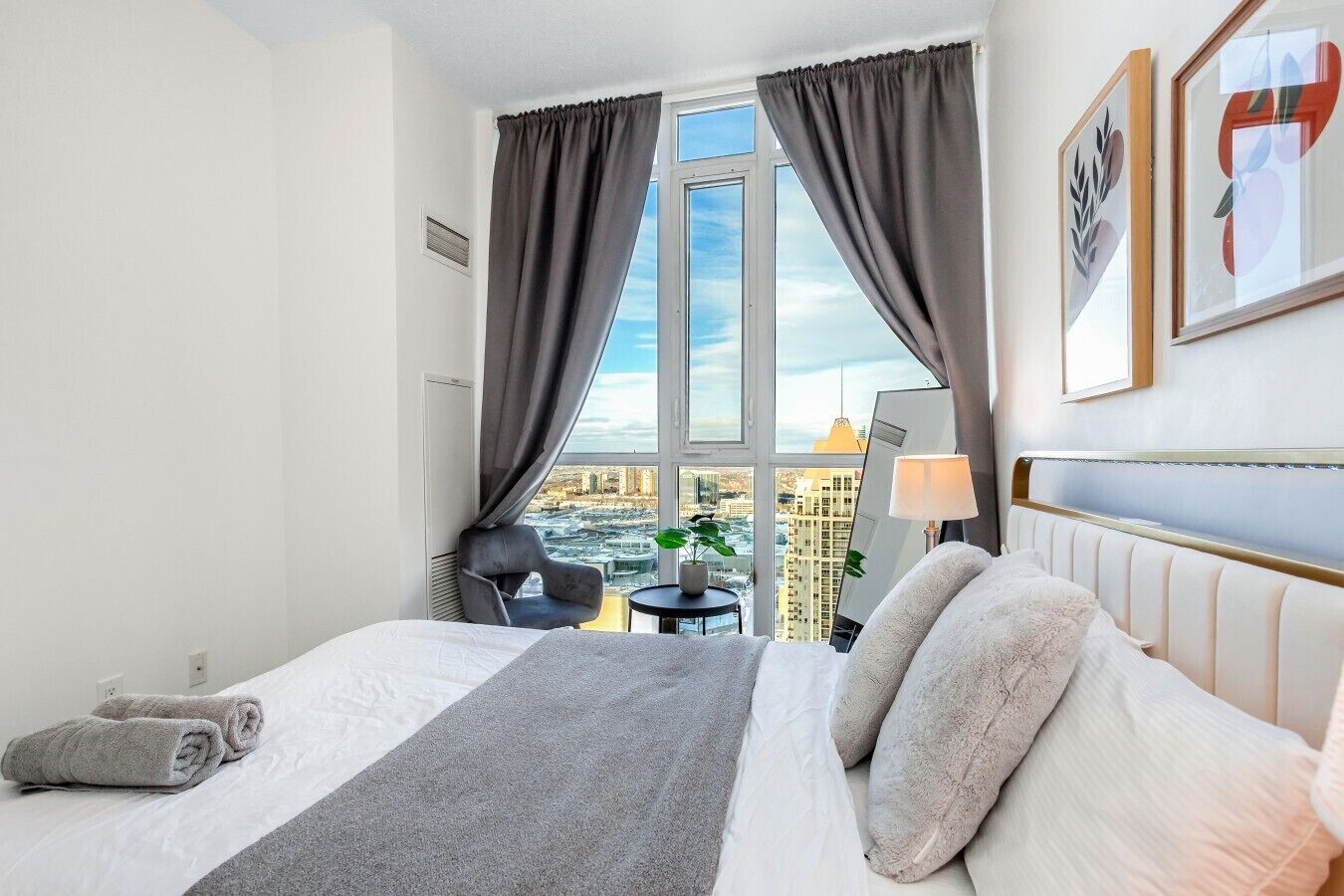 A hotel room with a bed, a chair, a small table, a lamp, and a view of the city through large windows.