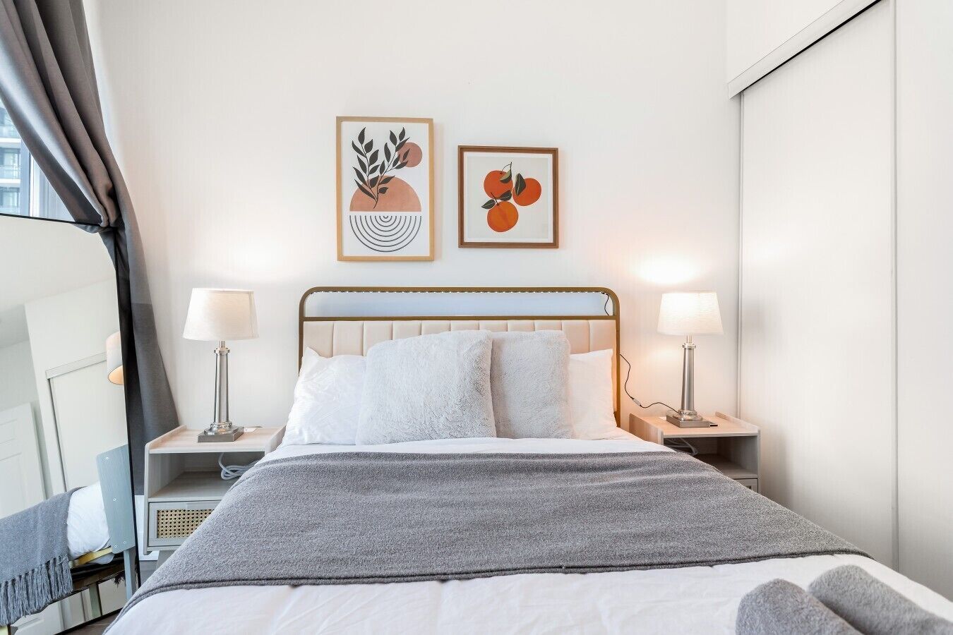 A hotel room with a bed, two bedside lamps, a gray throw blanket, and two framed artworks on the wall.