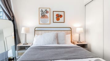 A hotel room with a bed, two bedside lamps, a gray throw blanket, and two framed artworks on the wall.