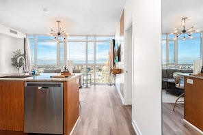 A modern kitchen with a dishwasher, a dining area with a view, and a living room with a sofa and a table. - Sky-High 2BR/2BA Condo | Best City Views+Parking (Mississauga)