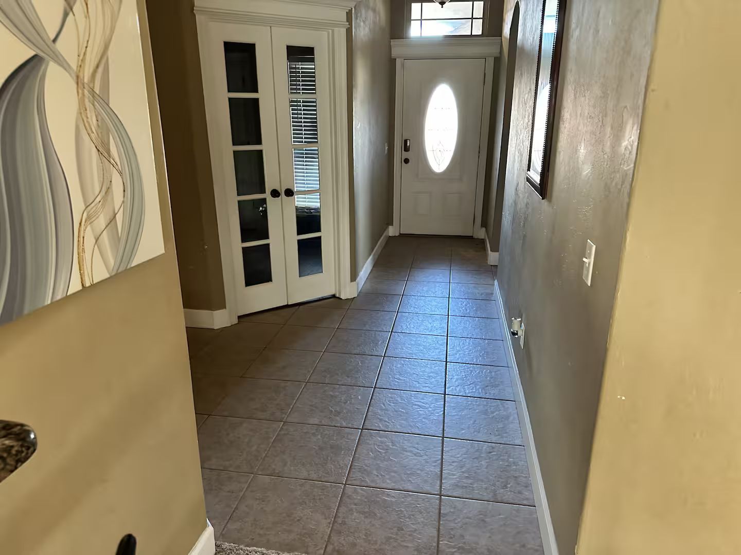 A hallway with a tiled floor, a white door with glass panels, and a wall with a partial view of a painting.