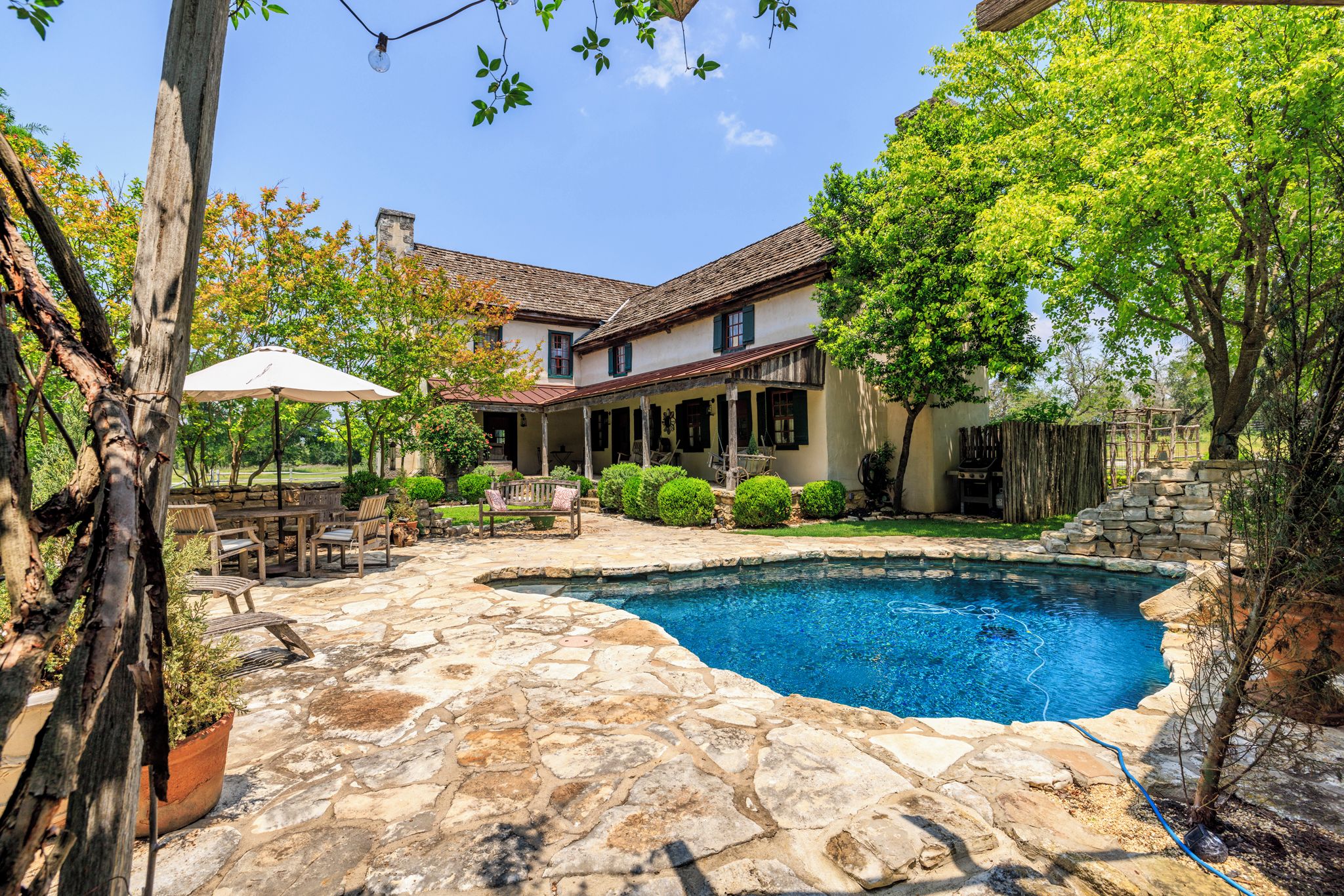 A stone patio with a swimming pool, outdoor furniture, and a house with a tiled roof.