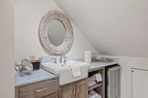 A bathroom with a white sink, a round mirror, and wooden cabinets. - A historic 3-story estate with a pool, sleeps 10 (Fredericksburg)