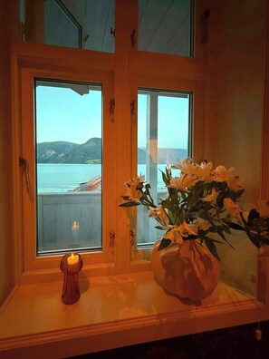 Una habitación con ventana que da a un cuerpo de agua y montañas, un florero con flores sobre una mesa, y una vela encendida.