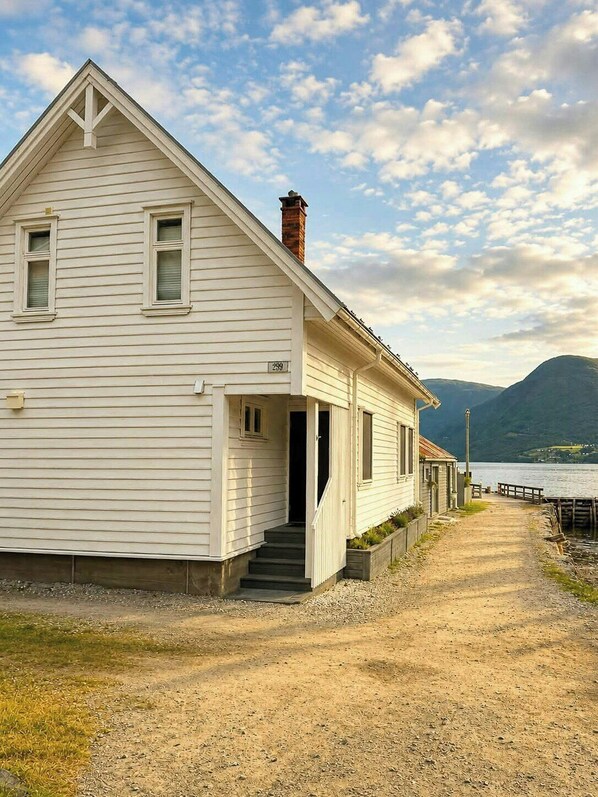 A white single-story house with a chimney, a number '299' on the side, a gravel driveway, and a body of water in the background.