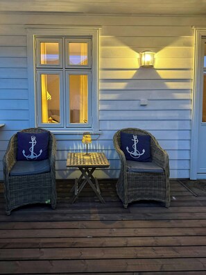 Two wicker chairs with anchor pillows, a small table with a lamp, and a window with curtains.