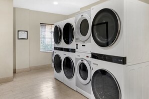 A laundry room with multiple washing machines and dryers, a framed sign on the wall, and a window with blinds.
