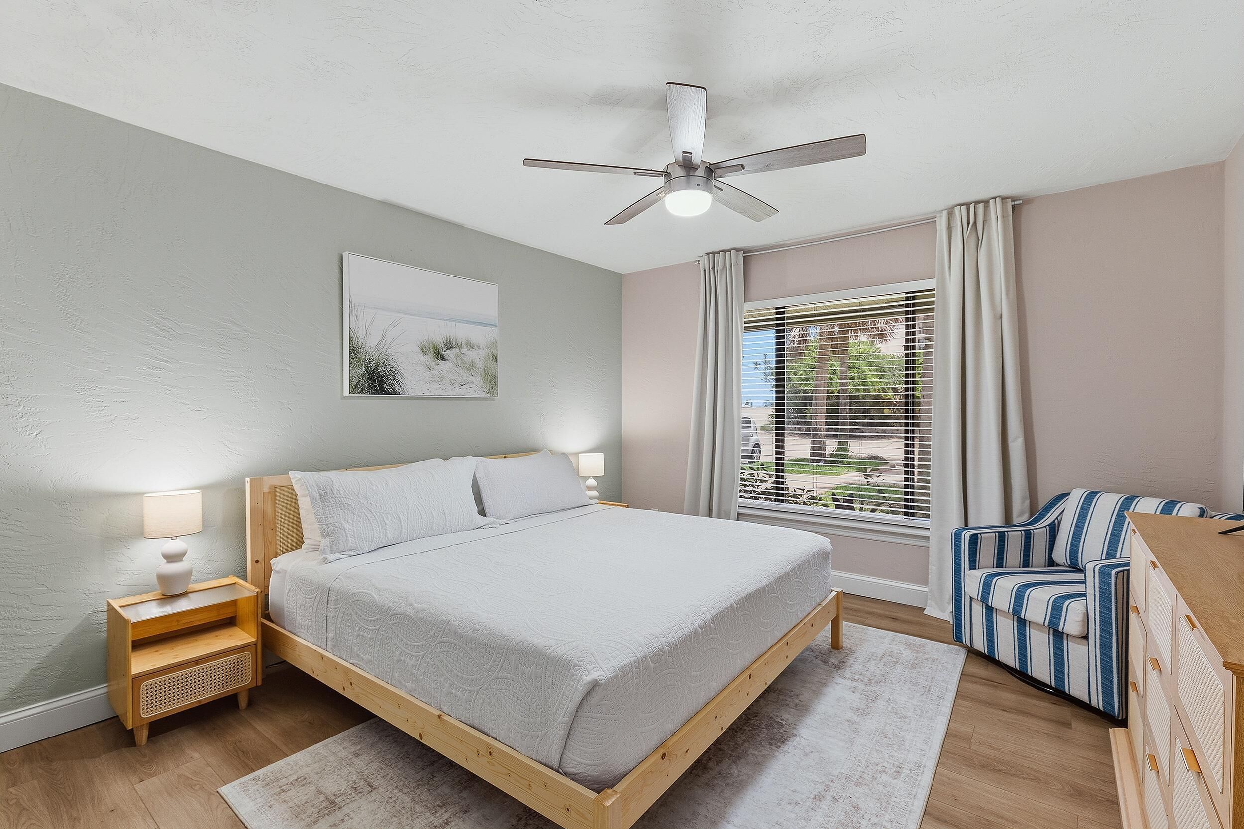 Un dormitorio con cama, mesitas de noche, ventilador de techo, ventana con cortinas y vistas al exterior.
