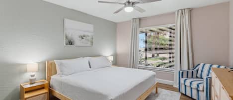 Un dormitorio con cama, mesitas de noche, ventilador de techo, ventana con cortinas y vistas al exterior.