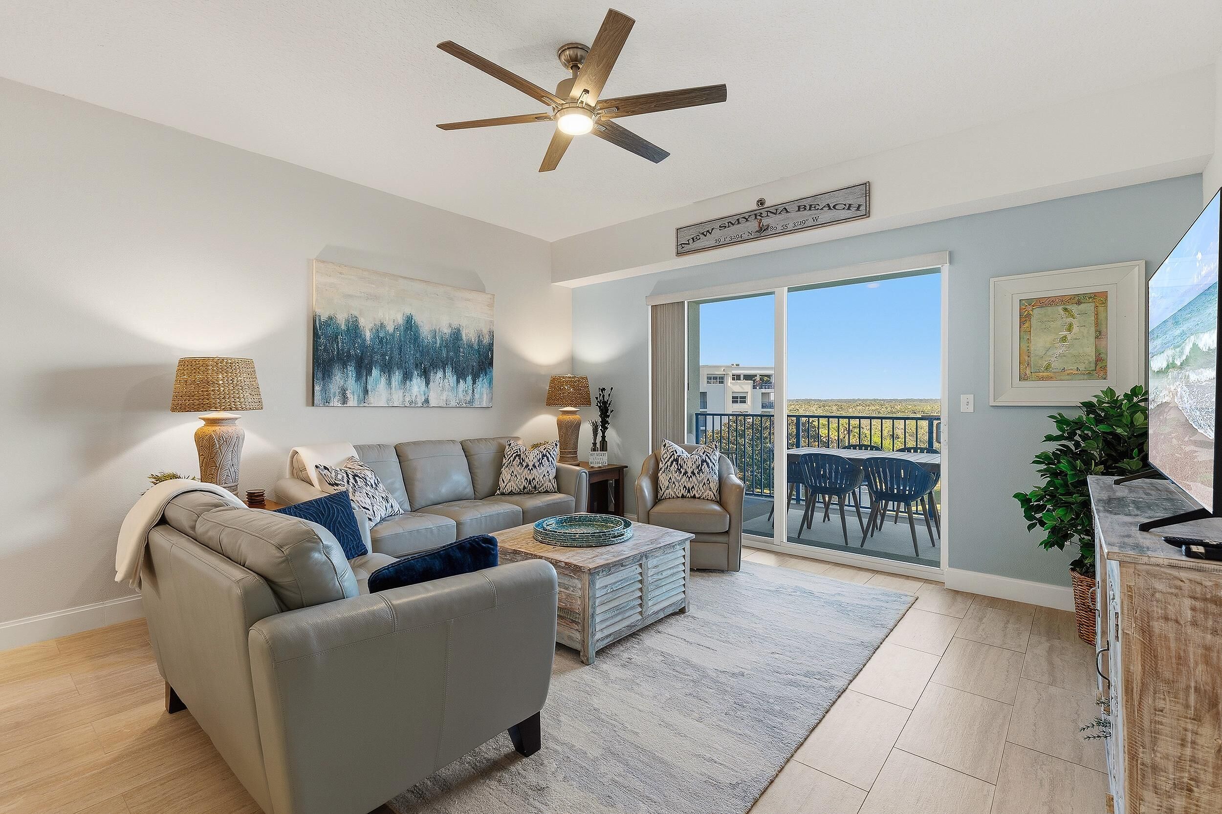 A modern living room with a grey sofa, a coffee table, and a ceiling fan. There is a large window with a view of the ocean and a balcony with outdoor furniture.
