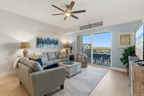 A modern living room with a grey sofa, a coffee table, and a ceiling fan. There is a large window with a view of the ocean and a balcony with outdoor furniture. - Oceanwalk 15-503 (New Smyrna Beach)