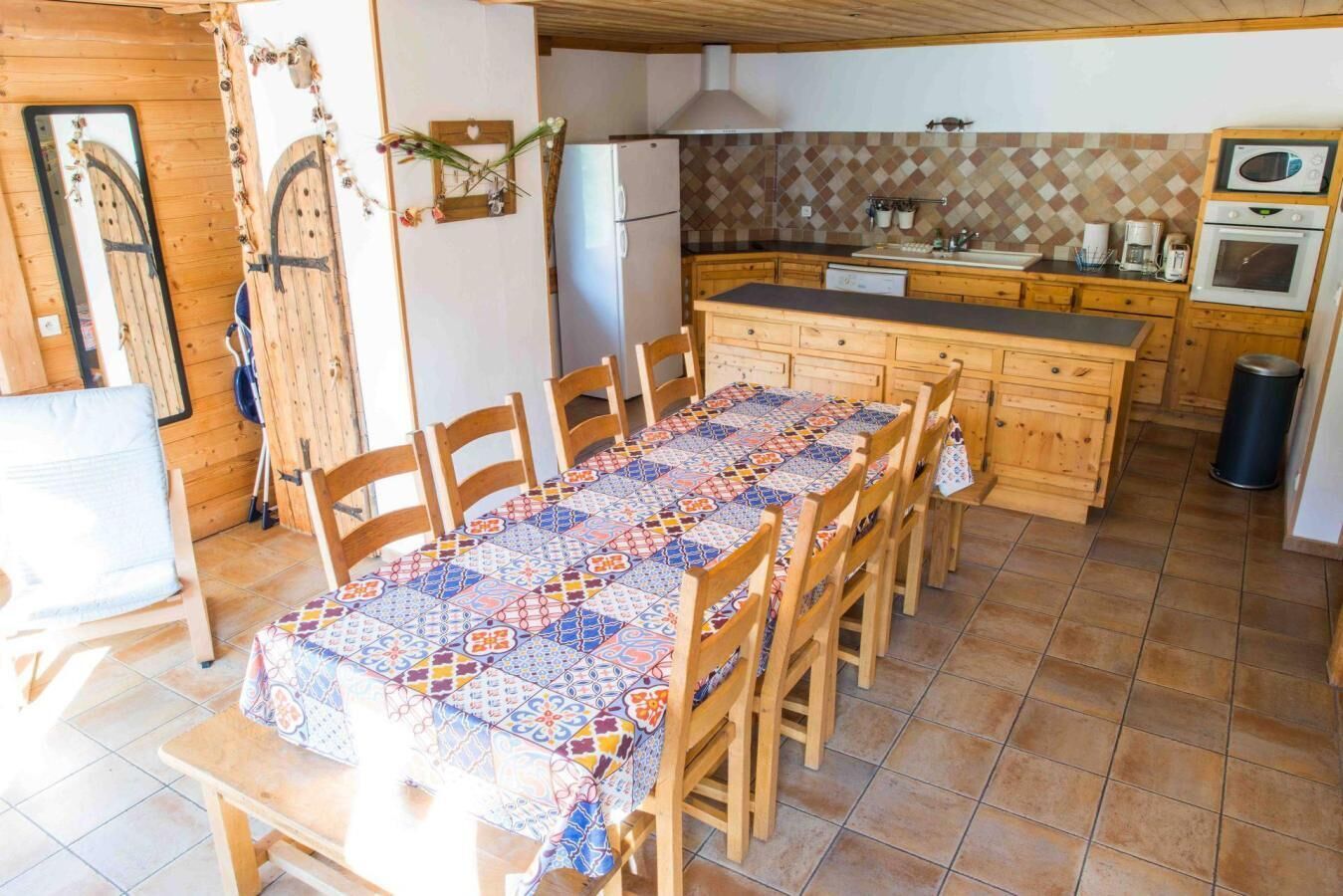 Un coin repas avec une longue table recouverte d’une nappe à motifs, des chaises en bois, une cuisine avec un mur recouvert de carreaux et un réfrigérateur.