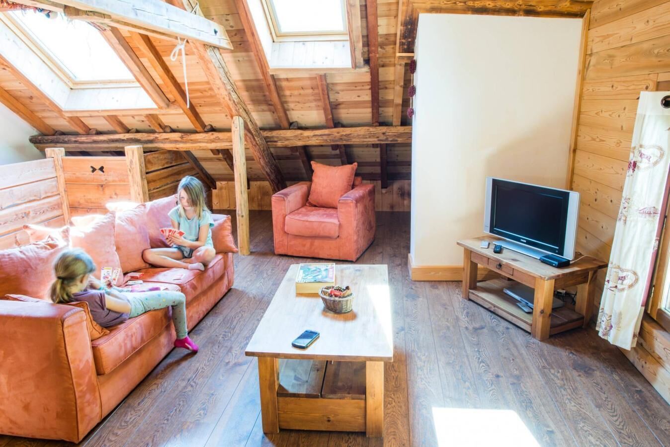 Een gezellige kamer met houten plafond, een televisie op een houten dressoir, een tafel met een mand en twee roze banken.