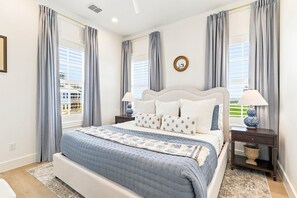 A bedroom with a large bed, two bedside tables, a nightstand, a window with curtains, and a wall-mounted mirror. - Seaside Cottage 101 YEA (Port Aransas)
