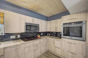 Une cuisine moderne avec des meubles de couleur bois clair, un pare-feu en carreaux noirs, des appareils en acier inoxydable et un plafond en bois.
