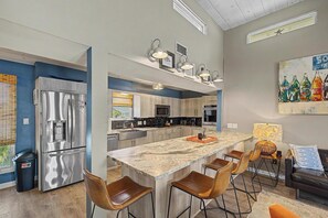 A modern kitchen with a central island, stainless steel appliances, and a dining area with bar stools. - 1112 Maldonado-Tropic Like Its Hot (Pensacola Beach)