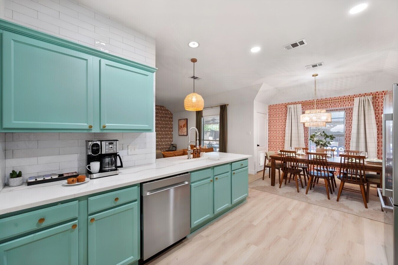 A modern kitchen with teal cabinets, a stainless steel dishwasher, and a dining area with a wooden table and chairs.