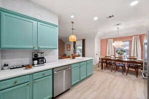 A modern kitchen with teal cabinets, a stainless steel dishwasher, and a dining area with a wooden table and chairs. - Hachie Hangout | Pool, Hot Tub, Pool Table, Fire Pit & Games (Waxahachie)