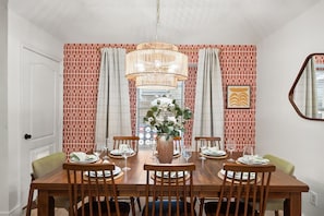 Une salle à manger avec une table en bois dressée pour six personnes, entourée d’un papier peint à motifs, d’un luminaire suspendu et d’un vase rempli de verdure.